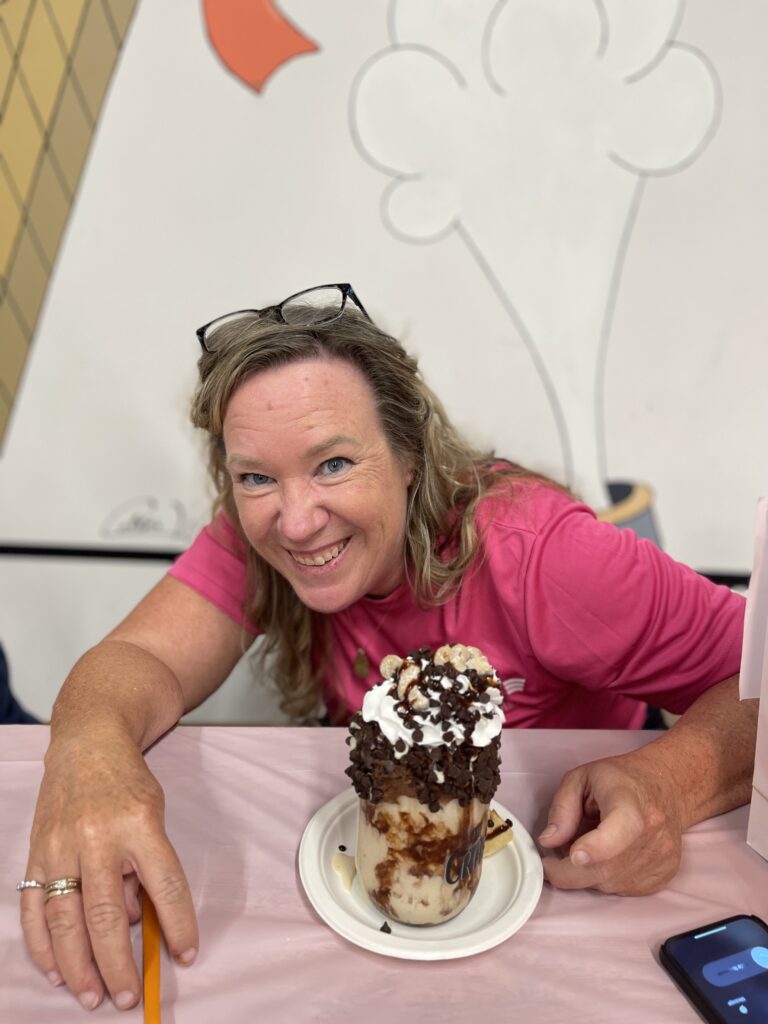 A woman wearing a pink shirt smiles while sitting in front of her on a table, holding a chocolate milkshake covered with toppings, during last year's Milkshakes & Bingo event.