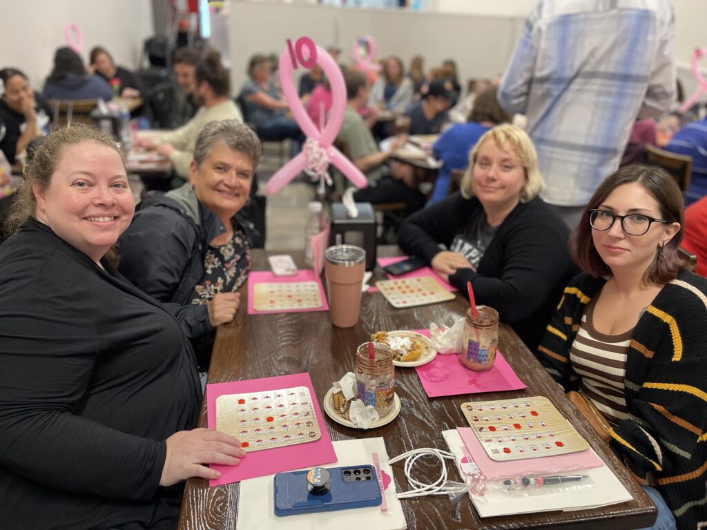 Four women are sitting at a table with bingo cards, milkshakes, and a pink ribbon balloon centerpiece during last year's Milkshakes & Bingo event.